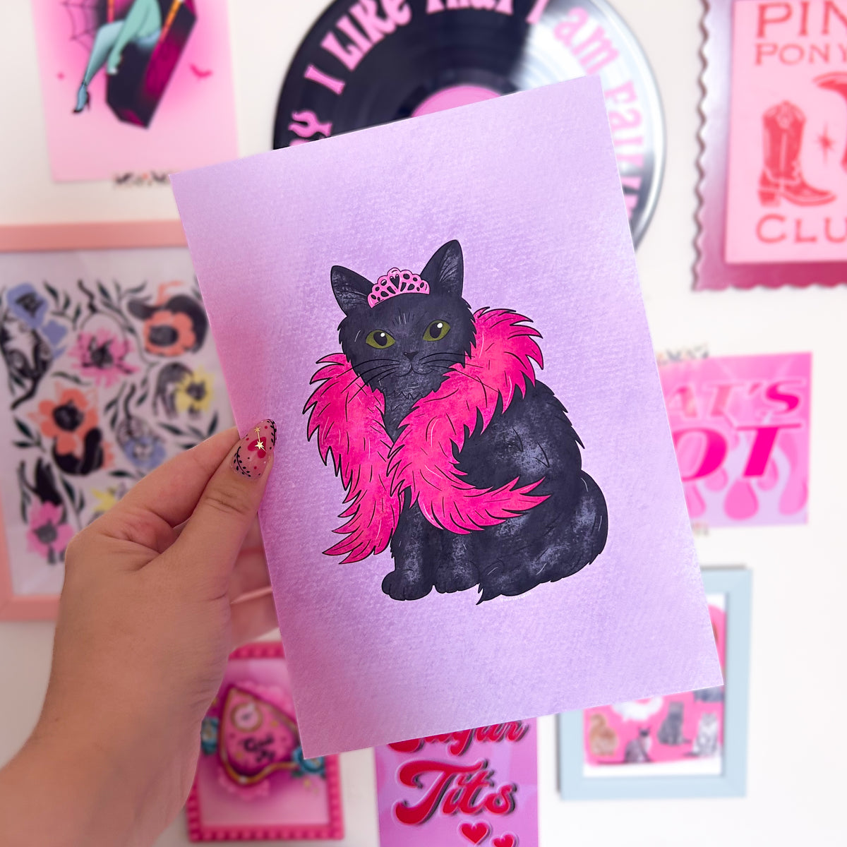 Print with a black cat illustration wearing a pink boa held by a hand against a colorful background.