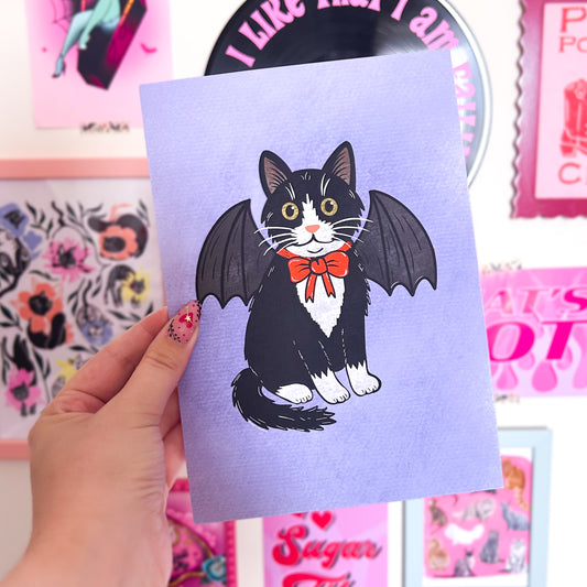 Print with a cat illustration wearing bat wings and a red bow tie held by a hand against a colorful background.