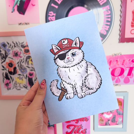 Print with a pirate cat illustration held by a hand against a colorful background.